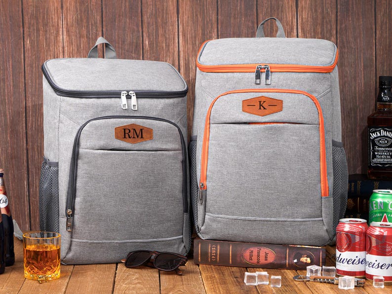 May include: Two gray insulated backpacks with personalized leather patches. One backpack has orange trim and the other has black trim. Both have a top handle and a front pocket. Sunglasses and a glass of amber liquid are in front of the backpacks.