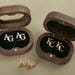 May include: Two wooden boxes with hinged lids, each containing a pair of cufflinks. The cufflinks in the left box are silver with the initials "AG" engraved on them. The cufflinks in the right box are gold with the initials "KC" engraved on them.