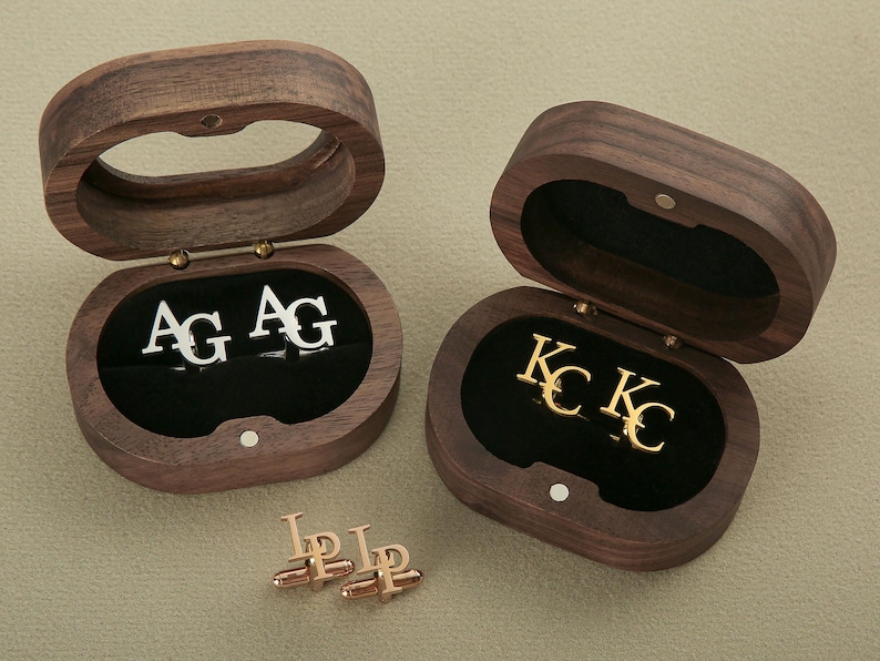 May include: Two wooden boxes with hinged lids, each containing a pair of cufflinks. The cufflinks in the left box are silver with the initials "AG" engraved on them. The cufflinks in the right box are gold with the initials "KC" engraved on them.