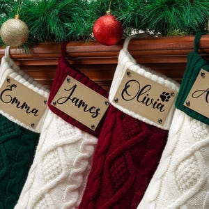May include: Four cable-knit Christmas stockings in dark green, white, and burgundy. Each stocking features a cream-colored cuff with a rectangular leather-like tag attached by four metal rivets. The tags are personalized with the names Emma, James, Olivia, and a partial name, with a paw print icon next to Olivia.