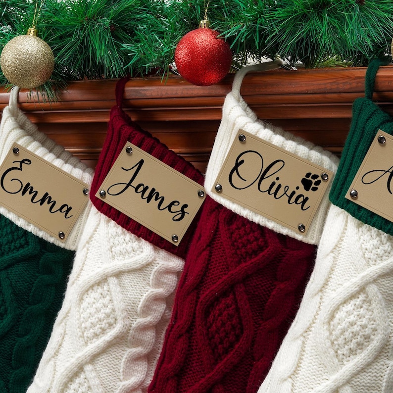 May include: Four cable-knit Christmas stockings in dark green, white, and burgundy. Each stocking features a cream-colored cuff with a rectangular leather-like tag attached by four metal rivets. The tags are personalized with the names Emma, James, Olivia, and a partial name, with a paw print icon next to Olivia.