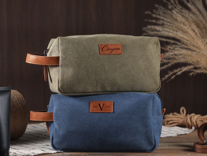 以下が含まれることがあります： 革の取っ手とジッパーが付いたキャンバス製の化粧ポーチが2つ。上のポーチは緑色で、茶色の革のパッチに「Ciayen」と書いてあります。下のポーチは青色で、茶色の革のパッチに「V」と書いてあります。
