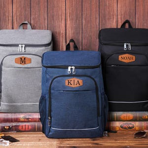 May include: Three personalized cooler backpacks in gray, navy blue, and black, each with a brown leatherette patch. The gray backpack has the letter "M", the navy blue backpack has "KIA", and the black backpack has "NOAH". The backpacks are on a wooden surface.