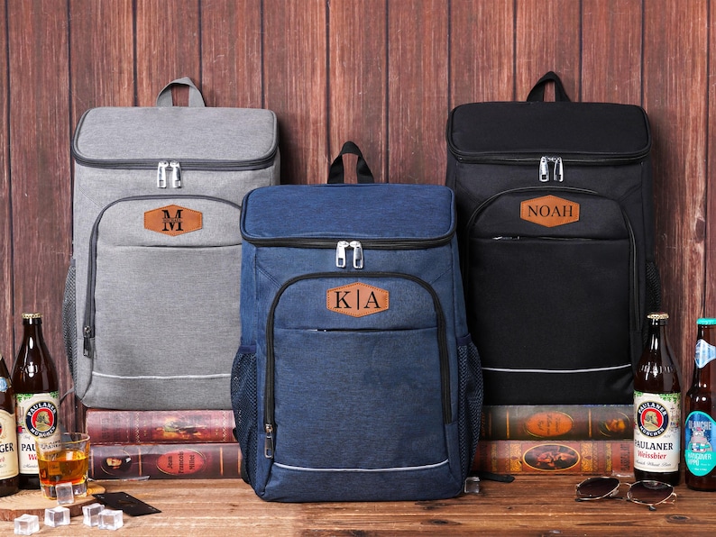 May include: Three personalized cooler backpacks in gray, navy blue, and black, each with a brown leatherette patch. The gray backpack has the letter "M", the navy blue backpack has "KIA", and the black backpack has "NOAH". The backpacks are on a wooden surface.