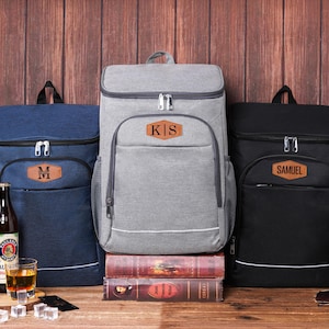 May include: Three insulated cooler backpacks in navy blue, gray, and black, each with a personalized leather patch. The navy backpack has an "M", the gray one "KIS", and the black one "SAMUEL". Beer bottles and a glass of amber liquid are in front.