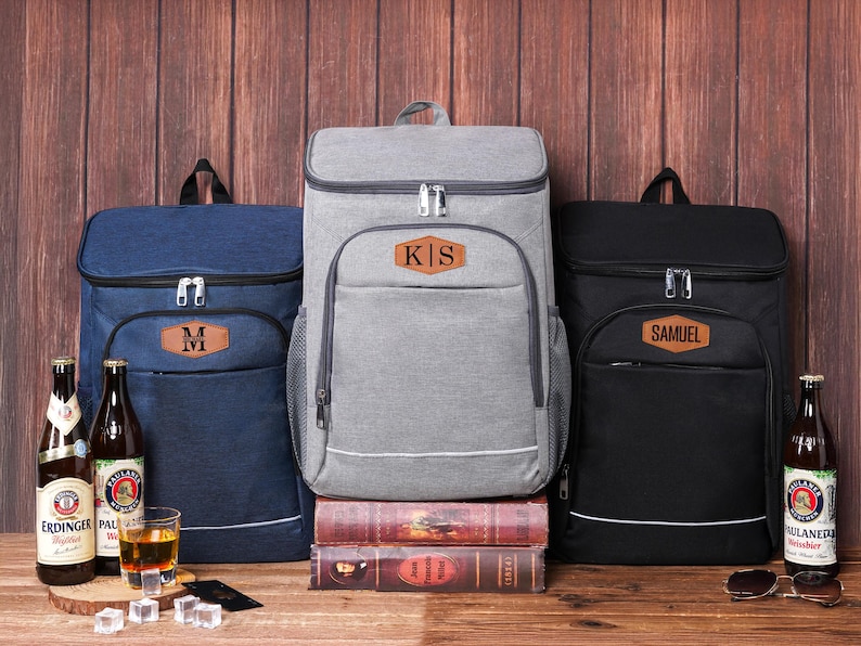 May include: Three insulated cooler backpacks in navy blue, gray, and black, each with a personalized leather patch. The navy backpack has an "M", the gray one "KIS", and the black one "SAMUEL". Beer bottles and a glass of amber liquid are in front.