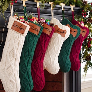 May include: A row of six cable-knit Christmas stockings in cream, dark green, and burgundy. Each stocking features a personalized leather patch with names including Evelyn, Olivia, James, Luna, Max, and a partial name, some shaped like bones.