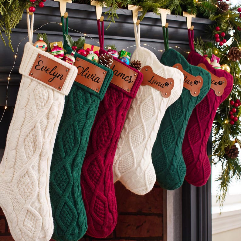 May include: A row of six cable-knit Christmas stockings in cream, dark green, and burgundy. Each stocking features a personalized leather patch with names including Evelyn, Olivia, James, Luna, Max, and a partial name, some shaped like bones.