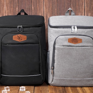 May include: Two insulated cooler backpacks, one black and one gray, with personalized leatherette patches. The black backpack has "NG" on its patch, while the gray one has "JAMES" and other text. Both have top handles and front pockets.