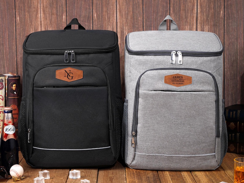 May include: Two insulated cooler backpacks, one black and one gray, with personalized leatherette patches. The black backpack has "NG" on its patch, while the gray one has "JAMES" and other text. Both have top handles and front pockets.