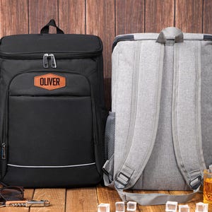 May include: Two insulated cooler backpacks. One is black with the name "Oliver" on a patch, the other is gray. Both have adjustable straps and mesh side pockets. Perfect for picnics or outdoor events.