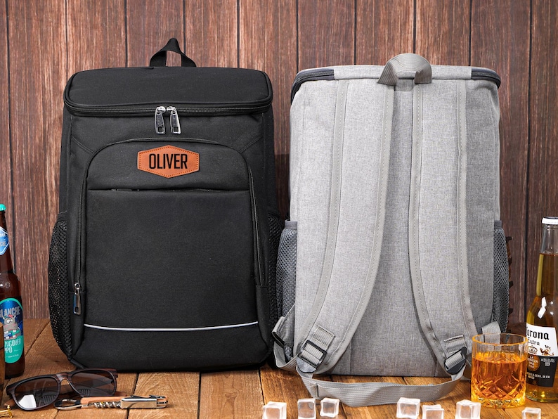 May include: Two insulated cooler backpacks. One is black with the name "Oliver" on a patch, the other is gray. Both have adjustable straps and mesh side pockets. Perfect for picnics or outdoor events.