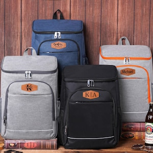 May include: A collection of insulated cooler backpacks in various colors: navy blue, gray, and black. Each backpack features a front pocket, a top handle, and a personalized leather patch. The backpacks are displayed with beer bottles and glasses.