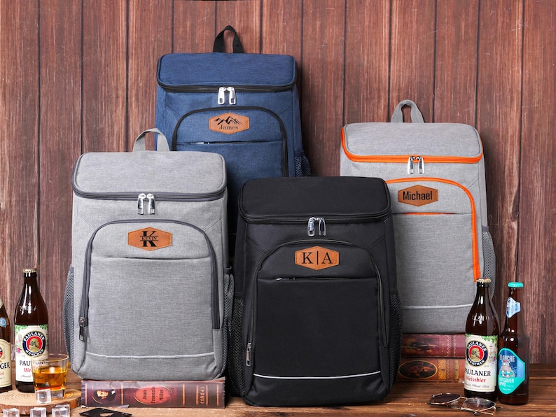May include: A collection of insulated cooler backpacks in various colors: navy blue, gray, and black. Each backpack features a front pocket, a top handle, and a personalized leather patch. The backpacks are displayed with beer bottles and glasses.