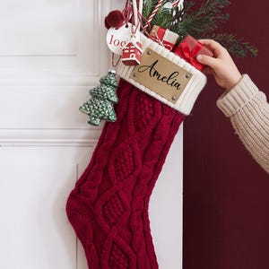 May include: A deep red cable-knit Christmas stocking with a white ribbed cuff featuring a tan leather patch engraved with the name Amelia. The stocking is filled with pine branches, small gift boxes, and a hanging green knit tree ornament.