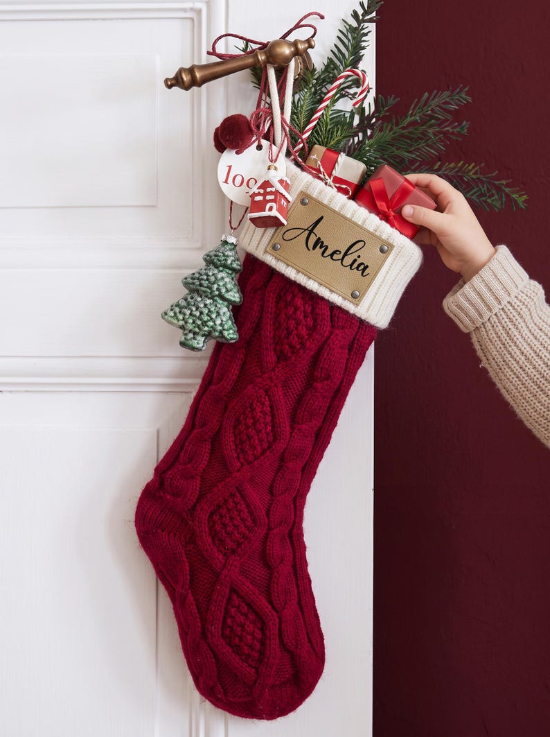 May include: A deep red cable-knit Christmas stocking with a white ribbed cuff featuring a tan leather patch engraved with the name Amelia. The stocking is filled with pine branches, small gift boxes, and a hanging green knit tree ornament.