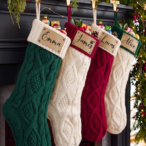 May include: Four cable-knit Christmas stockings in dark green, cream, burgundy, and cream. Each stocking features a personalized leather-look name tag with the names Emma, James, Lucas, and Alice, and a ribbed cuff. The stockings are filled with ornaments.