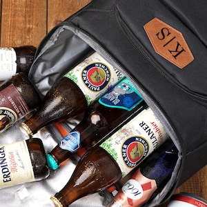 May include: A black insulated cooler bag filled with various beer bottles, including Erdinger, Paulaner, and Hoegaarden. The bag has a brown leather patch with the initials "K/S". The bottles are on a wooden surface.