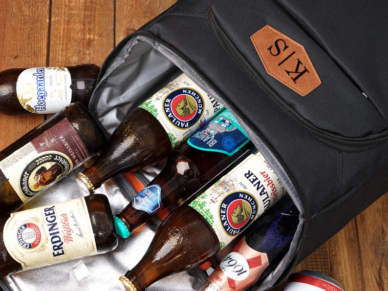 May include: A black insulated cooler bag filled with various beer bottles, including Erdinger, Paulaner, and Hoegaarden. The bag has a brown leather patch with the initials "K/S". The bottles are on a wooden surface.