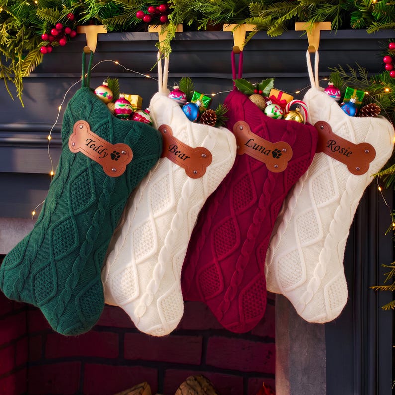 May include: Four cable-knit Christmas stockings in dark green, white, burgundy, and white. Each stocking features a brown leather bone-shaped tag with the names Teddy, Bear, Luna, and Rosie engraved, filled with small colorful holiday ornaments.
