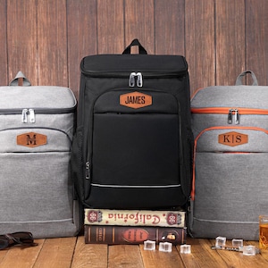 May include: A collection of personalized cooler backpacks in gray and black, with brown leatherette name patches. The backpacks are displayed on a wooden surface with drinks and accessories. The black backpack has the name "JAMES".