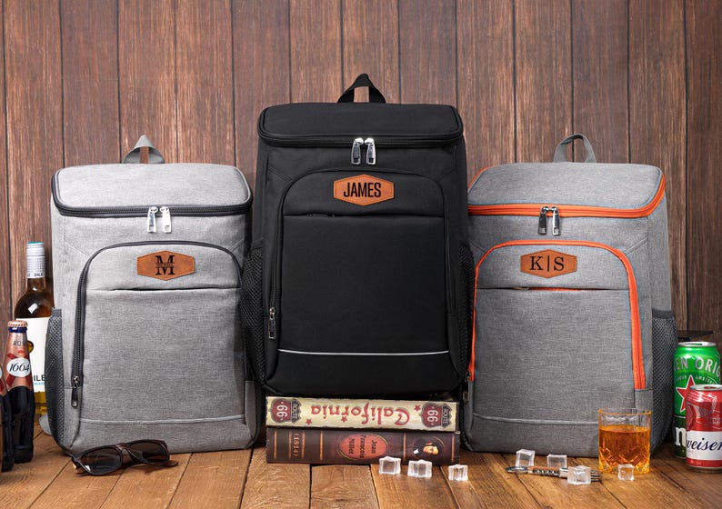 May include: A collection of personalized cooler backpacks in gray and black, with brown leatherette name patches. The backpacks are displayed on a wooden surface with drinks and accessories. The black backpack has the name "JAMES".
