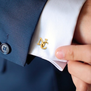 May include: Gold cufflinks with the letters "NC" intertwined. The cufflinks are worn on a white dress shirt.