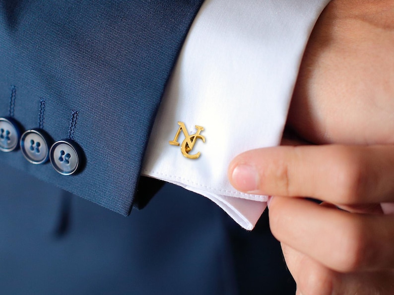 May include: Gold cufflinks with the letters "NC" intertwined. The cufflinks are worn on a white dress shirt.