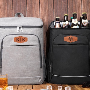 May include: Two insulated cooler backpacks, one gray and one black, with personalized leatherette patches. The gray backpack has the initials "KS", and the black one has "M". The black backpack is open, revealing several beer bottles.