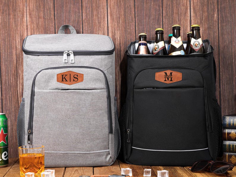 May include: Two insulated cooler backpacks, one gray and one black, with personalized leatherette patches. The gray backpack has the initials "KS", and the black one has "M". The black backpack is open, revealing several beer bottles.