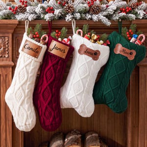 May include: Four cable-knit Christmas stockings in white, red, white, and dark green. Each features a leather-like name tag; two are rectangular with names Camila and James, and two are bone-shaped with names Luna and Max.