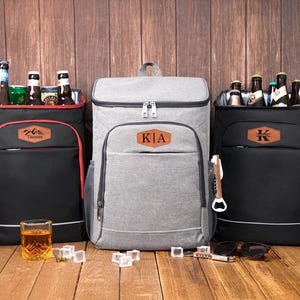 Może przedstawiać: Trzy izolowane plecaki chłodzące w kolorze czarnym i szarym, każdy z personalizowaną skórzaną naszywką. Plecaki są wypełnione butelkami i puszkami. Szklanka bursztynowego płynu i kostki lodu stoją z przodu.