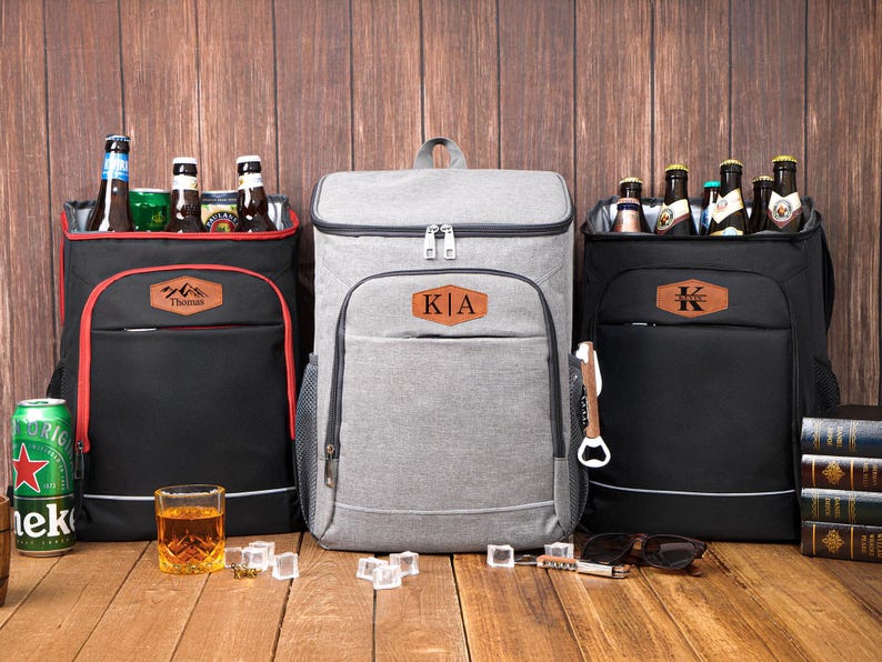May include: Three insulated cooler backpacks in black and gray, each with a front pocket and a personalized leather patch. The backpacks are filled with bottles and cans, perfect for outdoor events.