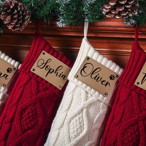 May include: Four knit Christmas stockings in alternating red and white colors, each featuring a tan rectangular leather-like patch secured with four metal rivets. The patches are personalized with the names Emma, Sophia, Oliver, and Noah in black script.