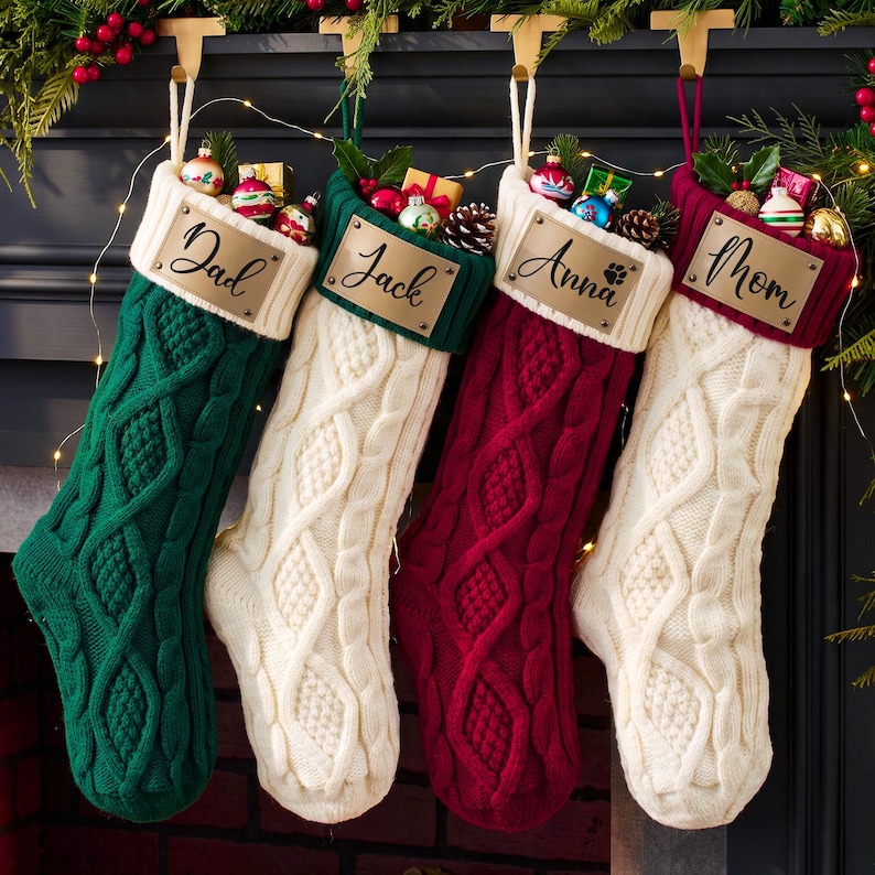 May include: Four cable-knit Christmas stockings in green, white, burgundy, and white. Each stocking features a cream-colored cuff with a rectangular leather-like tag engraved with the names Dad, Jack, Anna, and Mom, filled with small ornaments.