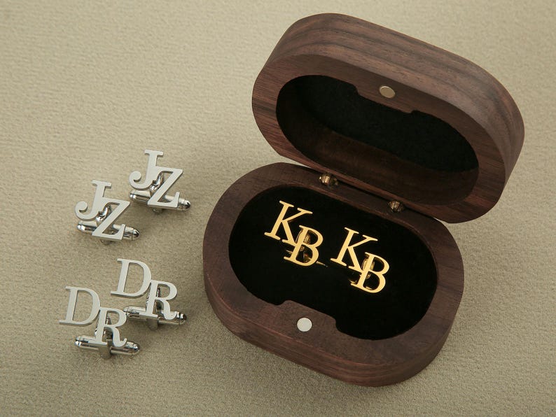 May include: A wooden box with a black velvet interior, containing two gold cufflinks engraved with the initials "KB".  The box is open, revealing the cufflinks.  Four silver cufflinks engraved with the initials "JZ" and "DR" are also shown.