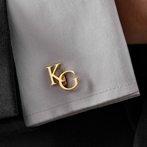 May include: Gold-colored cufflinks with the initials "KG" are displayed on a light gray dress shirt cuff. The cufflinks are in a serif font and are positioned on the cuff's edge. The background is a dark suit.