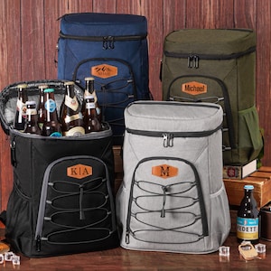 Puede incluir: Mochilas nevera aisladas en azul marino, negro, verde oliva y gris. Cada mochila tiene un parche de cuero personalizado con un nombre o iniciales. Una mochila está abierta, revelando botellas de cerveza.