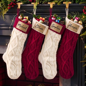May include: Four cable-knit Christmas stockings in alternating cream and deep red colors. Each stocking features a contrasting cuff and a rectangular metal nameplate engraved with the names Michael, Ethan, Sophia, and Harper respectively.