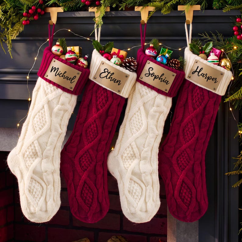 May include: Four cable-knit Christmas stockings in alternating cream and deep red colors. Each stocking features a contrasting cuff and a rectangular metal nameplate engraved with the names Michael, Ethan, Sophia, and Harper respectively.