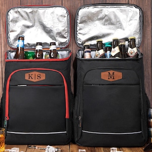 May include: Two black insulated cooler backpacks with red trim and personalized leather patches. One backpack has "KS" and the other has "M" on the patch. Both are open, revealing bottles of beer and other beverages.