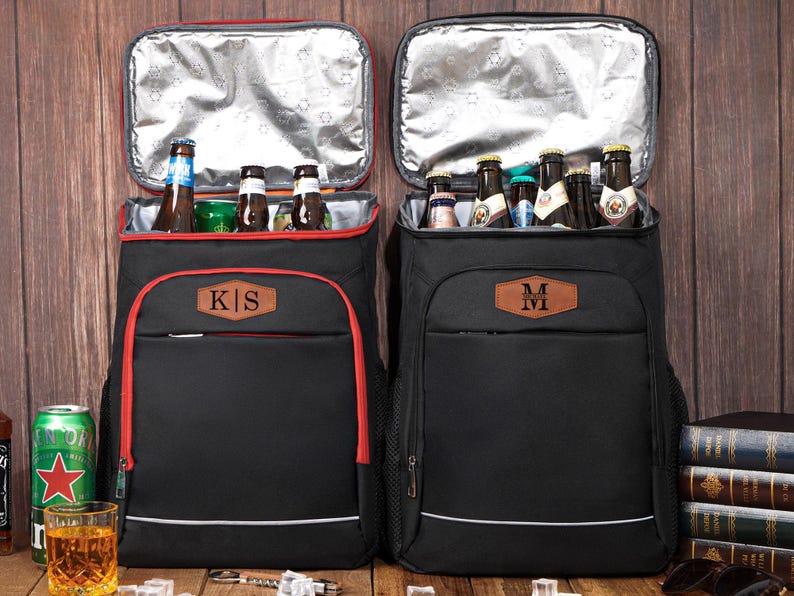 May include: Two black insulated cooler backpacks with red trim and personalized leather patches. One backpack has "KS" and the other has "M" on the patch. Both are open, revealing bottles of beer and other beverages.