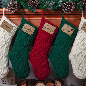 May include: Five cable-knit Christmas stockings in cream, green, and red hang from a mantel. Each stocking has a wooden name tag with names like "Greyson," "Kingston," and "Amelia." Pinecones and greenery decorate the mantel.