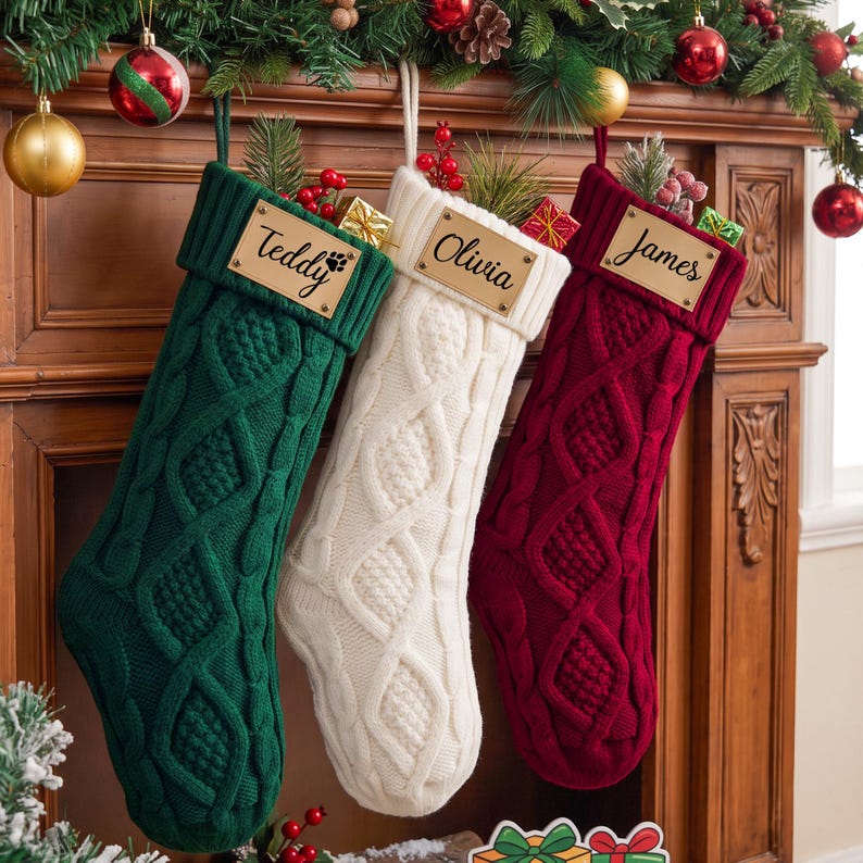 May include: Three cable-knit Christmas stockings in dark green, white, and deep red. Each stocking features a rectangular wooden name tag engraved with the names Teddy, Olivia, and James, and is filled with small pine sprigs and gift boxes.