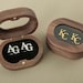 May include: Two wooden boxes with hinged lids. One box has a clear lid and contains two silver cufflinks with the initials "AG" engraved on them. The other box has a dark lid with the initials "KC" engraved in gold.