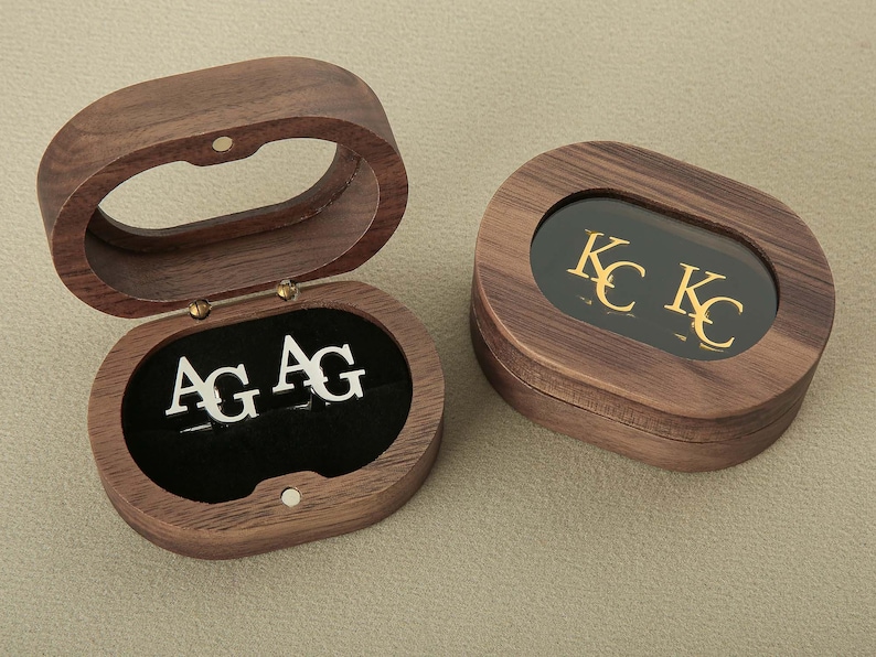 May include: Two wooden boxes with hinged lids. One box has a clear lid and contains two silver cufflinks with the initials "AG" engraved on them. The other box has a dark lid with the initials "KC" engraved in gold.