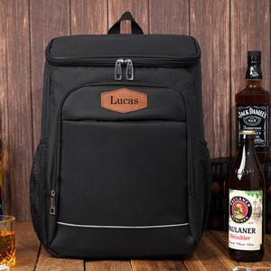 May include: Black insulated backpack with a top compartment and a front pocket. The backpack has a brown leather patch with the name "Lucas" on it. The backpack is next to bottles of beer and liquor.