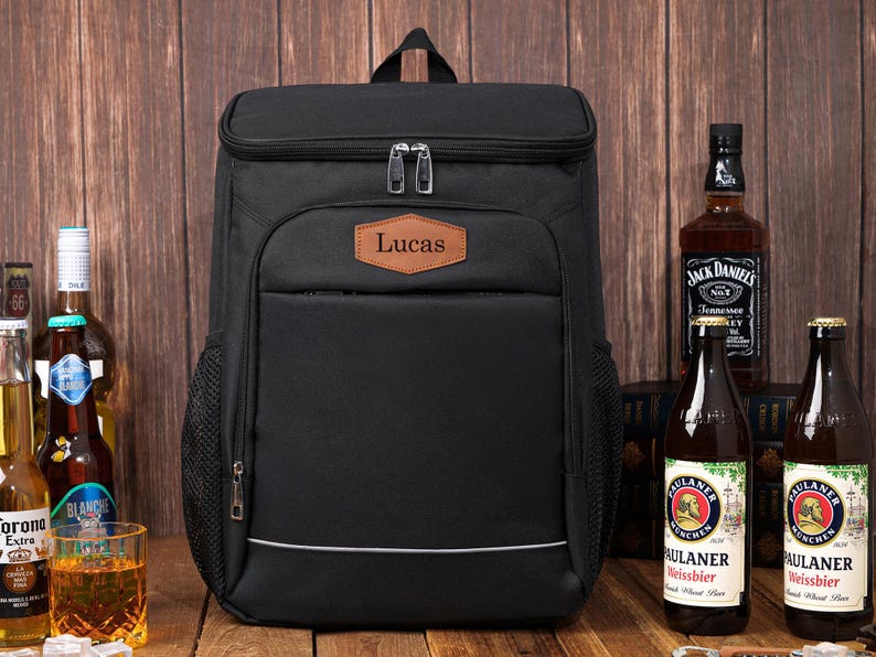 May include: Black insulated backpack with a top compartment and a front pocket. The backpack has a brown leather patch with the name "Lucas" on it. The backpack is next to bottles of beer and liquor.