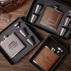 Pode incluir: Três conjuntos de frascos personalizados em caixas pretas, cada um com um frasco, copos de shot e um funil. Os frascos são de couro castanho, cinzento e bege, gravados com nomes e datas. Os copos de shot e os funis são prateados.