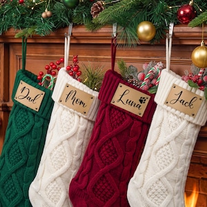 May include: Four knit Christmas stockings in green, white, burgundy, and white. Each stocking features a cable knit pattern and a rectangular leather-like tag attached with metal rivets. The tags are personalized with the names Dad, Mom, Luna, and Jack.